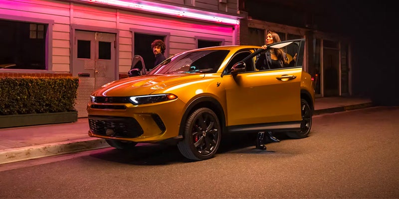 A golden Dodge Hornet with open passenger doors on a street at night, a man behind the driver and a woman exiting.