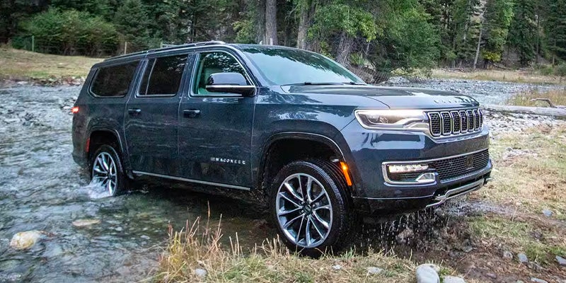 Dark gray Wagoneer SUV driving through a shallow, rocky stream with trees and brush in the background.