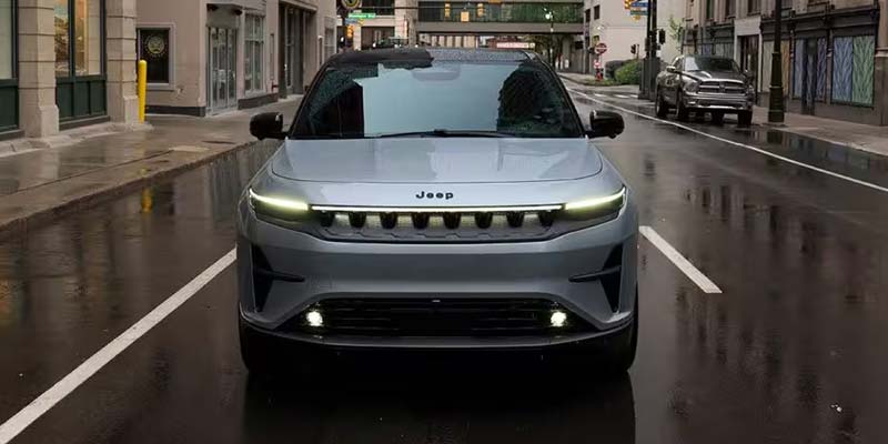 Front view of a silver-gray Jeep Recon EV on a wet city street with buildings and streetlights visible.