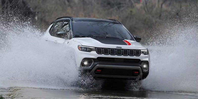 White Jeep Compass SUV driving through a puddle, splashing water high on both sides of the vehicle.