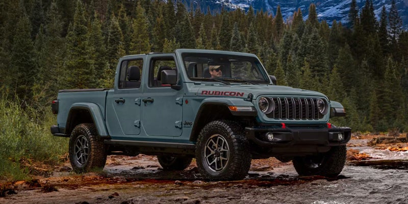 A light green Jeep Gladiator Rubicon pickup truck with a driver behind the wheel, parked on a muddy riverbank.