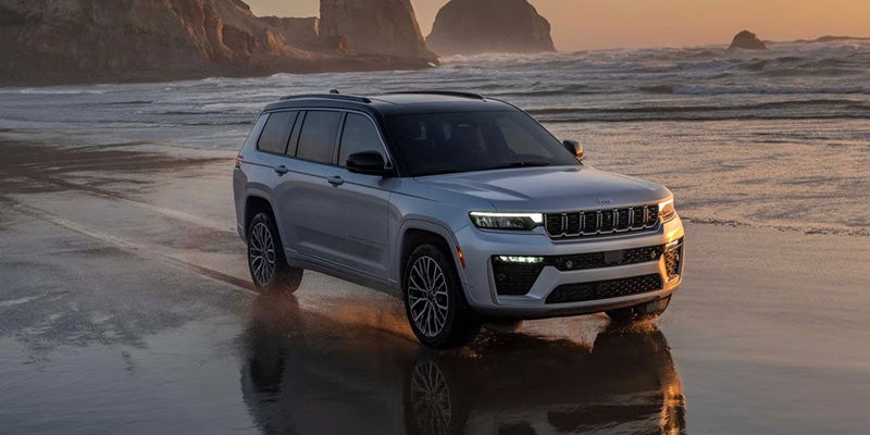 Silver Grand Cherokee L driving through shallow water on a beach at sunset, with ocean and rock formations behind it.