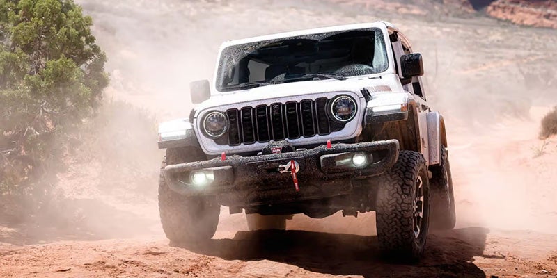 A white Jeep Wrangler kicks up dust while driving on a dirt road, trees, and rock formations in the background.