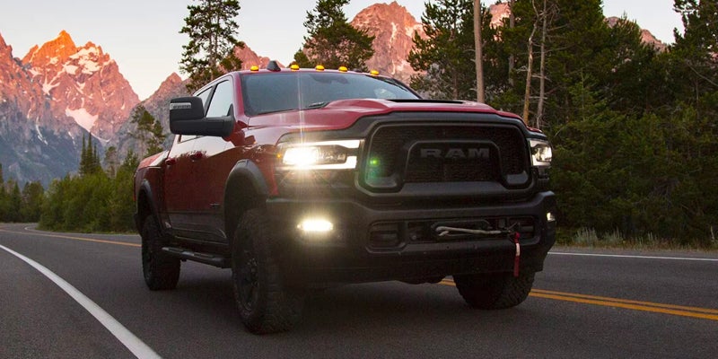 A powerful red off-road pickup truck with headlights on drives on a mountain road with sunlit peaks behind.