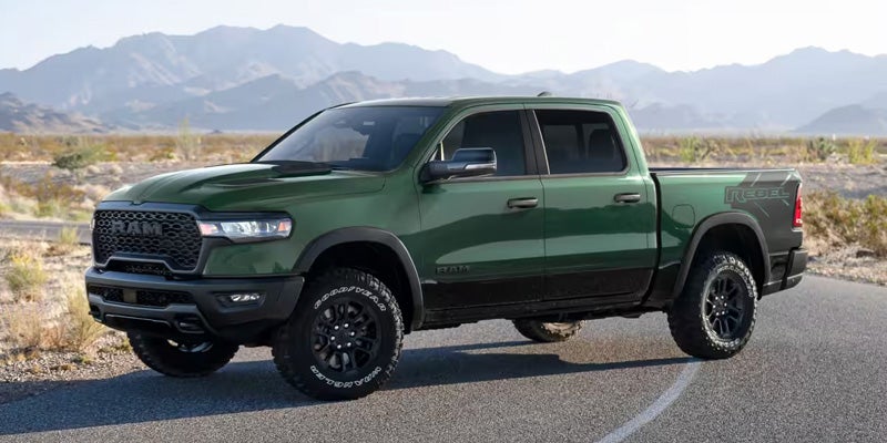 A dark green Ram Rebel pickup truck with black wheels, tinted windows, and a 'Rebel' decal on the bed, parked on an asphalt road with mountains and desert in the background.