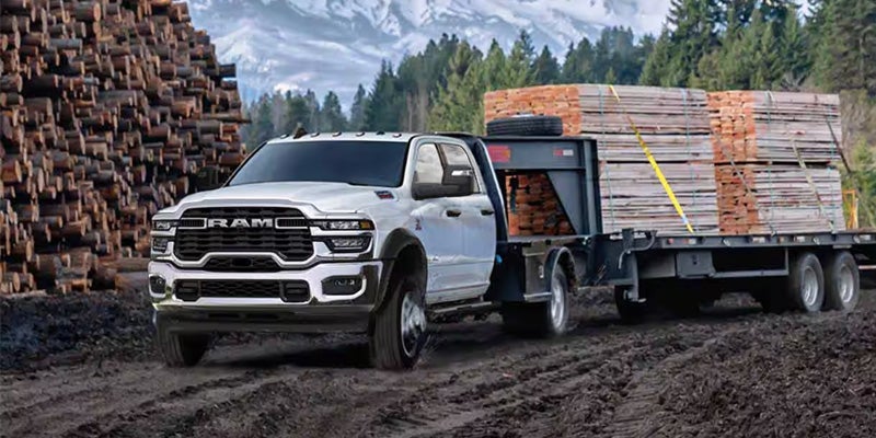A white Ram truck hauling a large flatbed trailer loaded with lumber on a muddy road, with a log pile and mountains in the background.