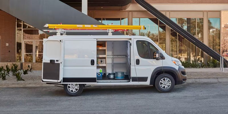 White cargo van with open side and rear doors, showing interior shelving and a yellow ladder rack on top.
