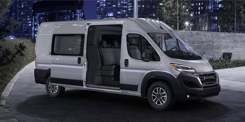 Silver Ram ProMaster van with the side door open, revealing three rows of dark gray seats, at night.